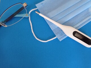 White electronic thermometer, glasses, medical mask  on a blue background. 