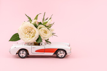 Retro white car delivering bouquet of white rose flowers on pink background. February 14, Valentine's day, 8 March, International Women's Day. Flower delivery. Copy space.