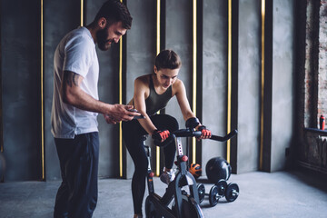 Determined young sportswoman spinning exercise bike with trainer nearby