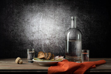 A bottle and two glasses, with a strong drink, and a white plate with a snack, a red napkin, on a wooden table top, on a background with a stain