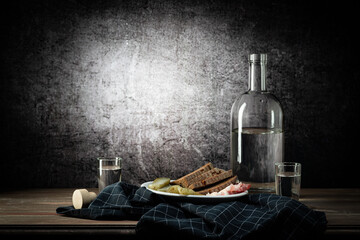 A bottle and two glasses, with a strong drink, and a white plate with a snack, a dark napkin, on a background with a stain
