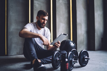 Positive caucasian sportsman in active wear spending time in gym using modern digital device for...