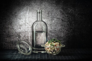 A bottle and two glasses, with a strong drink, and an olivier salad, on a dark tablecloth, on a background with a stain