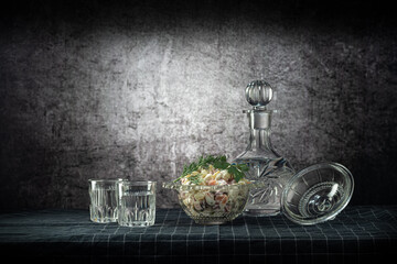 A decanter and two glasses, with a strong drink, and an olivier salad, on a dark tablecloth