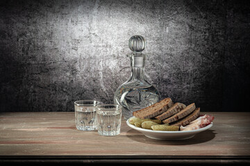 A decanter and two glasses, with a strong drink, with a white plate of snacks (pickles, lard, bread) on the wooden table top