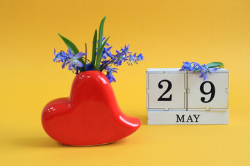 Calendar for May 29 : a bouquet in a heart-shaped vase with blue flowers and the number 29 on cubes, the name of the month of May in English, yellow background