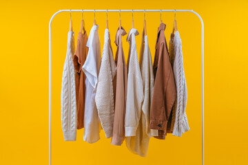 Variety of warm cozy light-colored female clothing hanging on white rack, isolated on yellow background