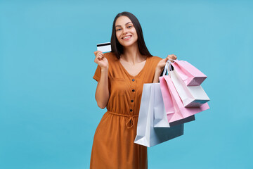  Studio shot of beautiful happy smiling brunette girl posing with pile of shopping bags and credit card in hands, isolated on pastel blue background