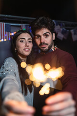 Pareja joven con bengalas de chispas abrazadas,sonriendo y besandose con una caravana de fondo con al bandera de estados unidos