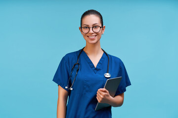 Studio portrait of smiling young brunette doctor woman in surgical uniform and trendy eyeglasses, posing over light blue background with tablet computer in hand