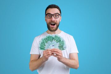 Portrait of happy excited young bearded man holding ward of euro banknotes in hands unable to belive in his victory in online lottery - obrazy, fototapety, plakaty