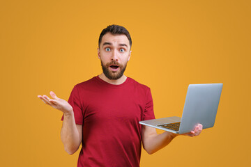 Young bearded man holding laptop computer in hand and looking directly at the camera with confused face expression