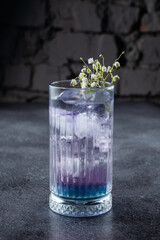 A cocktail in a tall glass with blue liqueur and ice, decorated with white flowers on gray background.