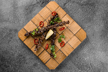Grilled chicken hearts with lime on wooden skewer, garnished with chili, cilantro, sesame on gray background.