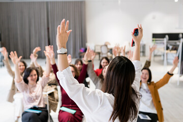 Motivation training for women. Rear view of speaker with group of people raising their hands up at...