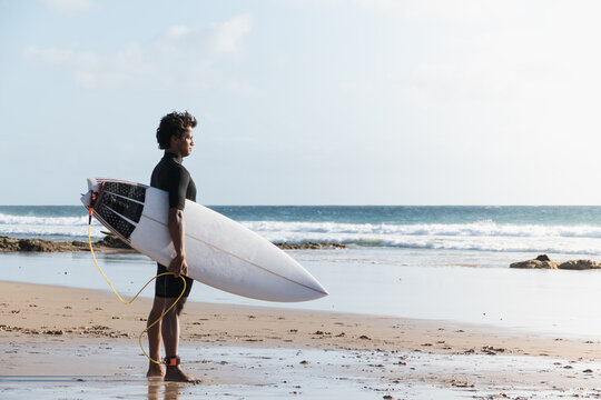 Young african malagasy surfer standing at the beach shore watching the waves holding surfboard