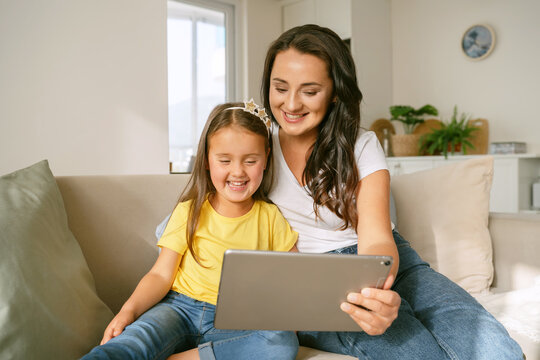 Little Girl Daughter And Positive Young Mother Watching Funny Videos Together On Digital Tablet While Relaxing On Sofa In Living Room At Home And Enjoying Family Weekend. Kids And Modern Technologies