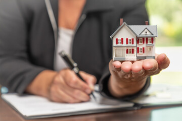 Woman signing real estate contract papers holding small model home in front.