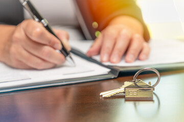 Woman signing real estate contract papers with house keys and home keychain in front.