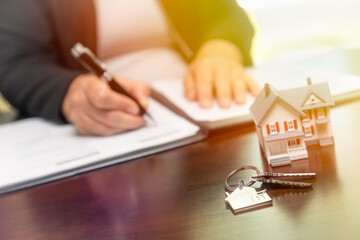 Woman signing real estate contract papers with house keys, home keychain and small model home in...