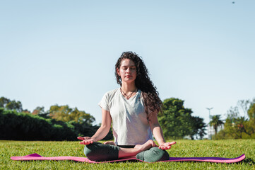 beautiful latin girl, meditating in lotus flower pose, on a pink mat and with the sky in the background, in a park, yoga and lifestyle concept.