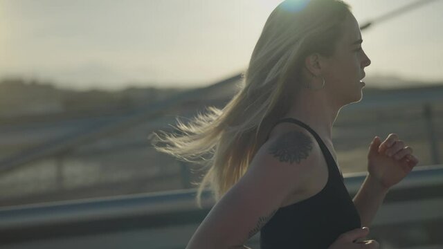 Close-up tracking shot of a pretty young woman running in the city at the seaside at sunset. Girl works out for wellbeing in summer.  