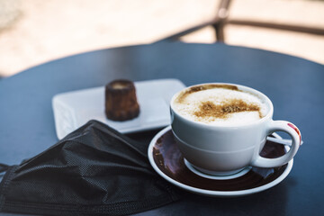 Cup of coffee cappuccino or latte and face mask on a table. Measures in outdoor cafe, bar or restaurant after reopening. Reduction of quarantine restrictions.