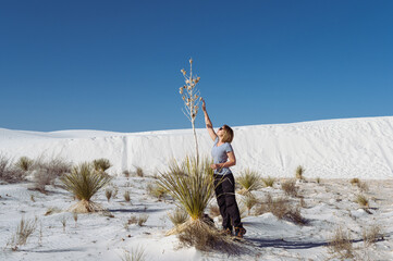 a woman trying to reach with her hand  the top of the yucca tree over the background of blue sky and white sands dune