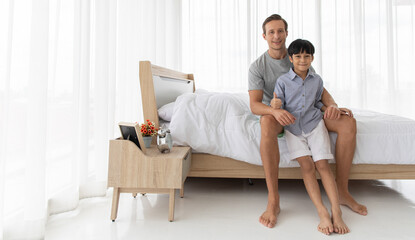 Father sitting on bed in ome bedroom and hugging his mixed-race son with a happy and joyful pose....