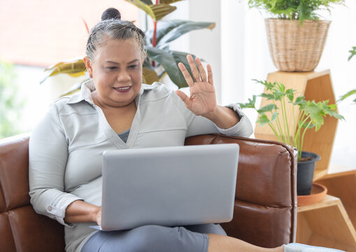 An Older Senior Asian Woman Sitting On Sofa And Wave Hand To Notebook Labtop Computer In Living Room. Idea For Older People Learning New Skill Technology.