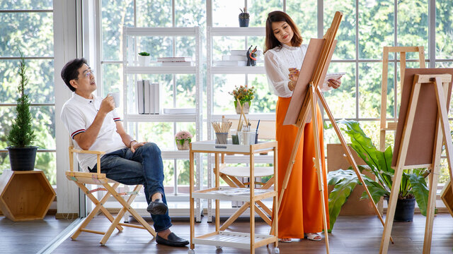 .Senior woman artist painting image in studio at home with green small tree in little garden and full of natural light. Her older husband sitting and drinking coffee and watching for her work