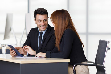 Caucasian happy handsome successful businessman in black formal suit smiling listening to Asian pretty businesswoman who use pencil pointing at tablet screen explaining important business information