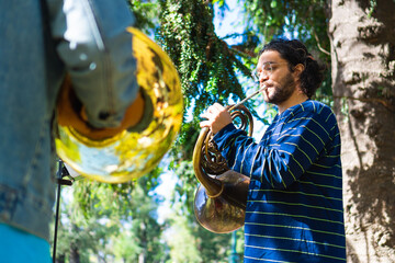 latin man playing the french horn in a park rehearsing with his band. low angle view