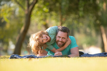 Fototapeta premium Weekend activity happy family lifestyle concept. Dad and child embracing and hugging together outdoors. Father and son laying on grass in summer park spending time outdoor.