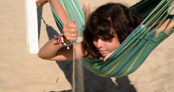12 years old brunette girl laying in a hammock and playing with sand at the beach, 4k slow motion