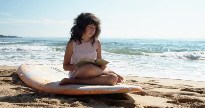 A cute girl is sitting on a surfboard on the beach and reading a book during summer vacation. 4k slow motion