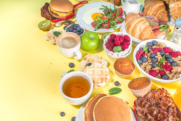 Various traditional breakfast food - fried eggs with bacon, muesli, oats, waffles, pancakes, burger, croissants, fruit berry, coffee, tea and orange juice, yellow table background copy space top view