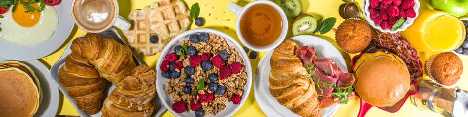 Various traditional breakfast food - fried eggs with bacon, muesli, oats, waffles, pancakes, burger, croissants, fruit berry, coffee, tea and orange juice, yellow table background copy space top view