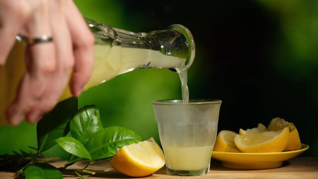 Limoncello is a traditional Italian liquor, alcoholic drink made of lemons. Female hand pours limoncello from a decanter into a chilled shot glass, fresh lemons on background.