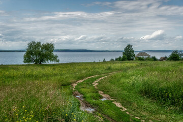 View of the bank of the Kama River (Russia) on a cloudy spring day. A dirt dirt road turns picturesquely to a single wooden house with a vegetable garden. textured white-gray clouds, fresh greens 