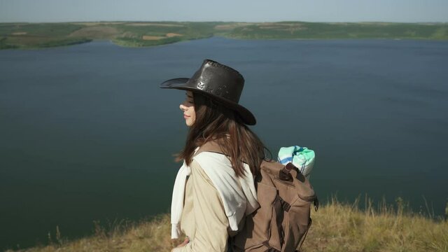 Positive woman in casual wear and cowboy hat standing on edge of hill and looking at wide Dniester river. Female hiker with backpack having active time during weekends.