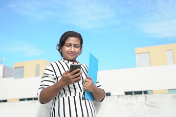 Asian businesswoman holding a file and reading her cellphone outside her office. Outdoor setting.