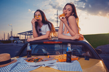 Charming females are eating pizza while posing in yellow car cabriolet with french fries and soda...