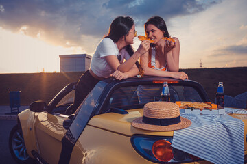 Two young females eating pizza, smiling, posing in yellow car with french fries, hat and soda in glass bottle on its trunk. Fast food. Mock up