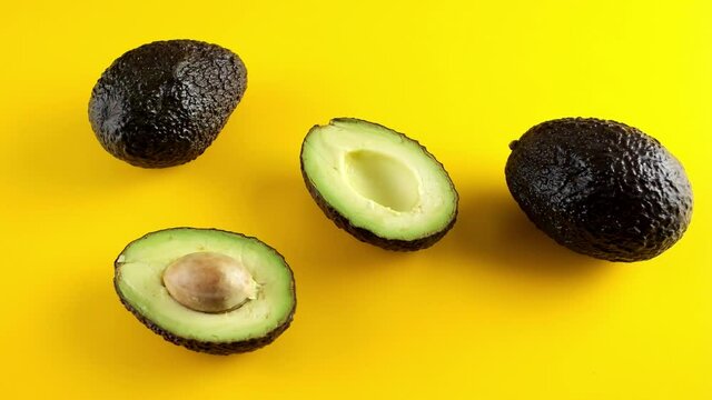 Halved avocado with a stone and two whole ripe avocados on yellow background, 4K UHD dolly side pan