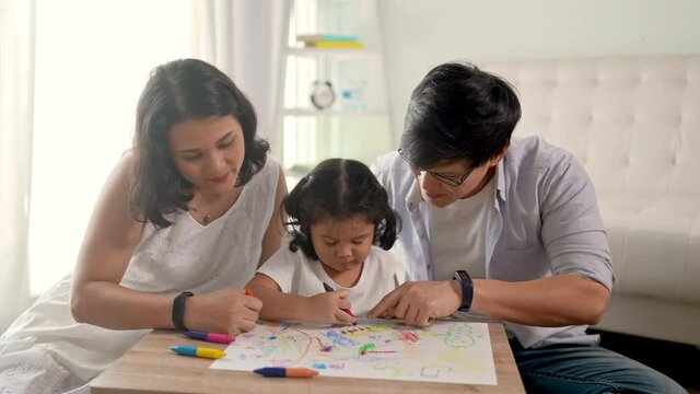 Happy Asian family at home. Parents teach little daughter using color pencil drawing on paper book in living room. Father and mother with cute child girl kid having fun leisure homeschooling together