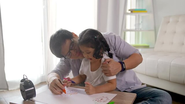 Happy Asian family at home. Father teach little daughter using color pencil drawing on paper book in living room. Dad and child girl kid having fun leisure activity and homeschooling together at home