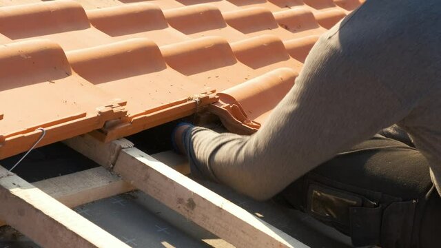 Closeup of worker hands installing yellow ceramic roofing tiles mounted on wooden boards covering residential building roof under construction.