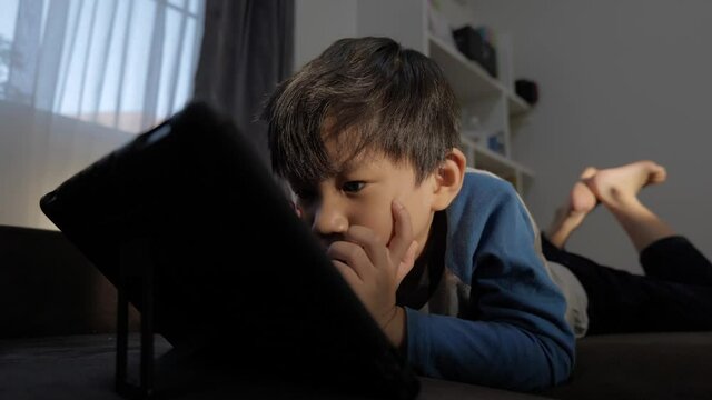 Asian boy watching computer tablet with addict expression on the couch. A problem for the kid who keeps using the screen.