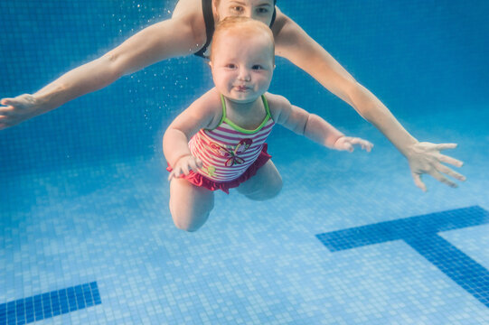 Mom with daughter are immersed in water, swimming under water in paddling pool. Diving baby. Learning infant child to swim. Young mother or swimming instructor and happy little girl.